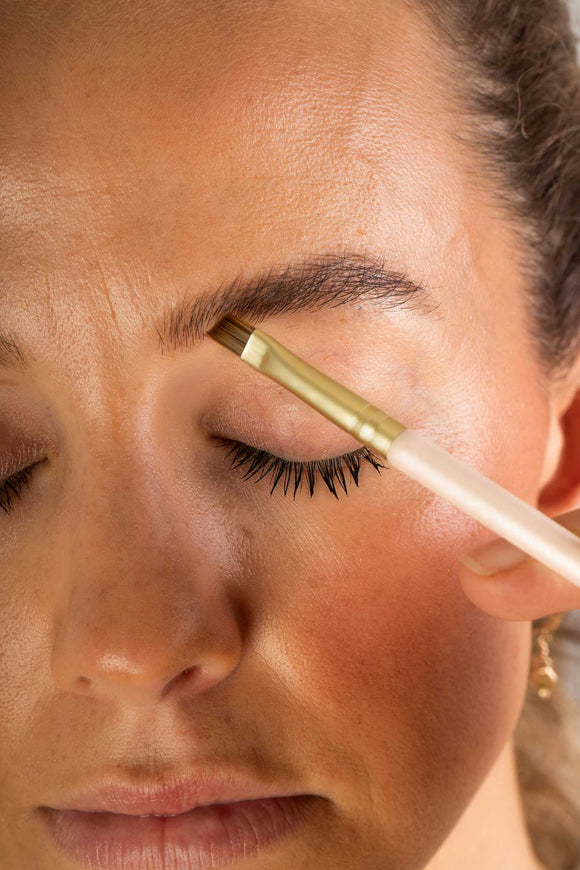 Model is applying the brow balm using an angled brush in her eyebrows in the shade Coffee.