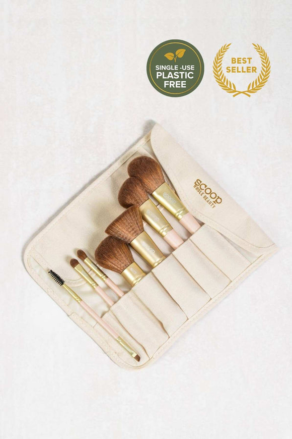 7 piece vegan brush set with beige canvas roll up bag. Brushes have a wooden pink handle and medium-dark brown bristles. Brushes include brow brush, two eyeshadow brushes, a kabuki brush, foundation brush, angled brush and blusher brush.