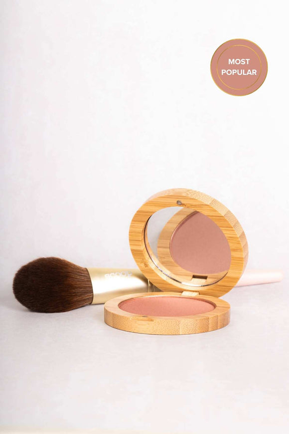 bamboo clamshell style round compact with pink blusher inside one side and a mirror on the inside of the lid. behind it is a blusher brush with a gold and pink handle with a soft looking dark brown fading bristles.